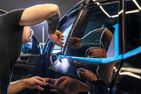 A tattooed man applies protective film to a black car at an auto detailing studio