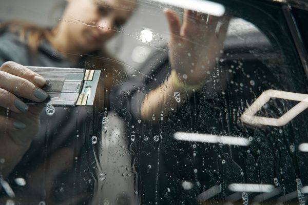 Experienced Caucasian service station worker sliding scraper over clear windscreen protection film