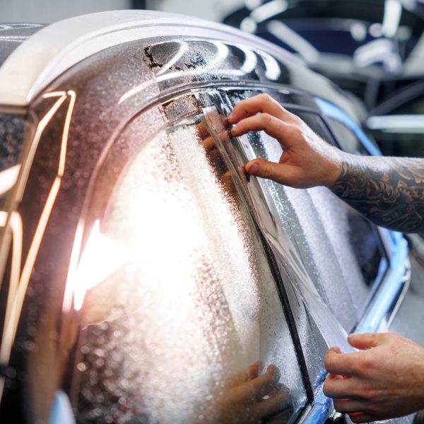 Sticker of a protective film on the headlight of a car in a car detailing studio. Man uses a knife with a blade, close-up of his hand